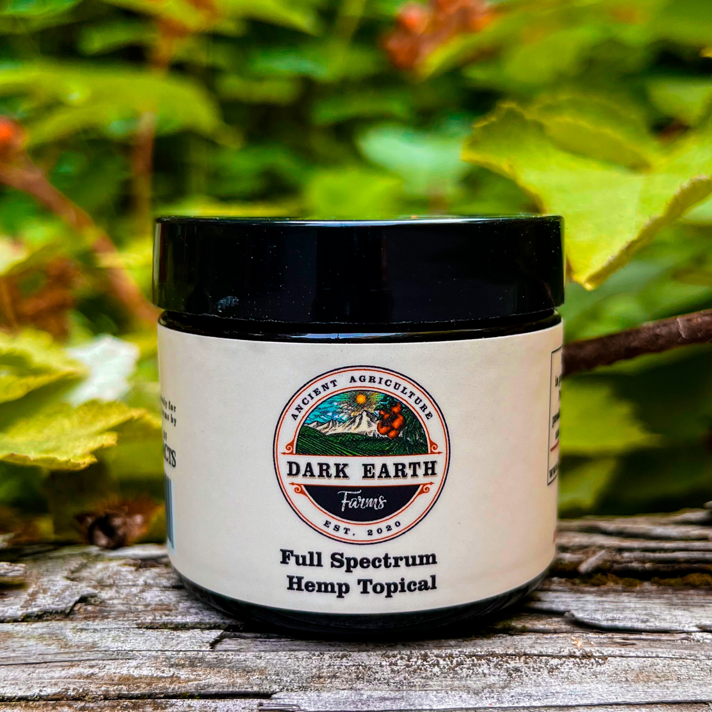 An image of a small jar labeled Dark Earth Farms Full Spectrum Hemp Topical, featuring a blue and yellow label with the brand name and product information. The jar contains 2000mg of CBD cream with terpenes and cannabinoids, designed for targeted relief from pain and inflammation. The jar is placed on a wooden surface, surrounded by hemp leaves, and the cream is shown oozing out of the jar, ready for application.